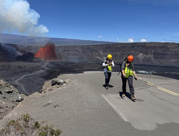 color photo of two scientists walking away after observing the lava fountain in the distance. 