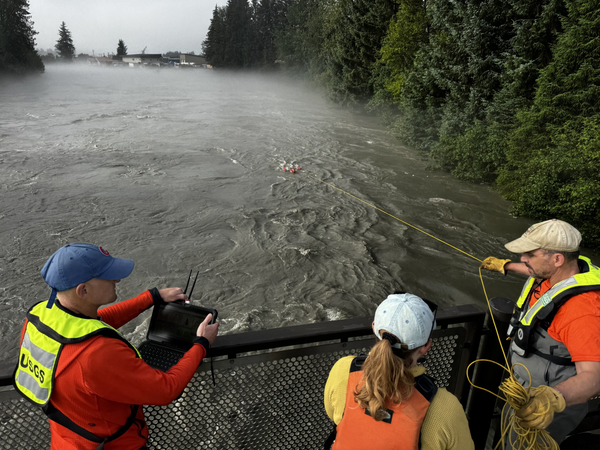 Three scientists stand on a bridge above a flooded river holding a rope attached to scientific equipment in the water below. 