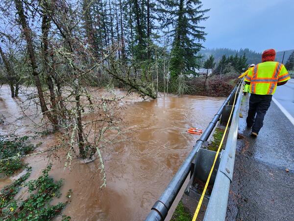 Swollen brown river water flowing through downed trees. Two technicians in safety gear pull ADCP across the channel 