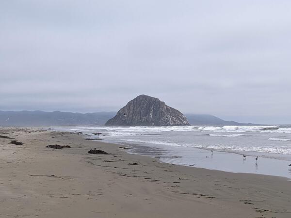 An isolated, large rocky hill rises above a foggy Pacific beach lined with crashing waves
