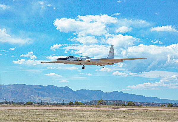 A white aircraft descends with landing gear extended set against a backdrop of blue sky, scattered clouds, and mountains
