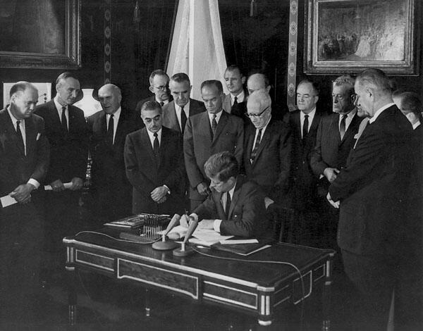 Black and white photo of President John F. Kennedy surrounded by people in suits signing a document