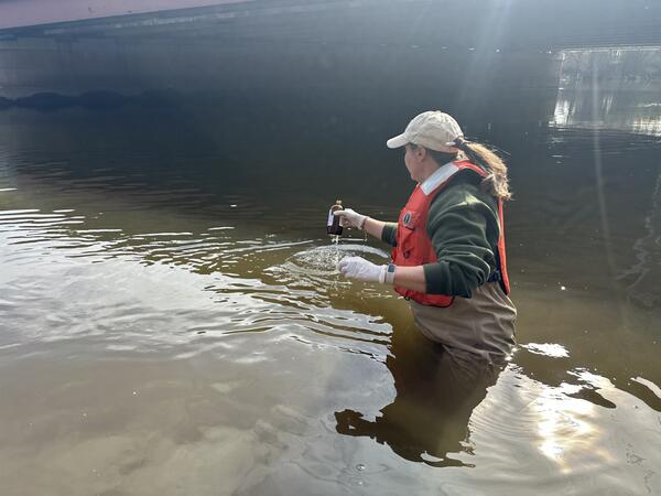 Woman with long brown hair in a ponytail and ballcap wearing a floatation vest wading into a river lifting a small bottle