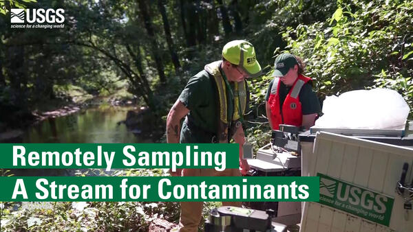 Two people lift heavy equipment adjacent to a stream. Text reads: Remotely Sampling A Stream for Contaminants.