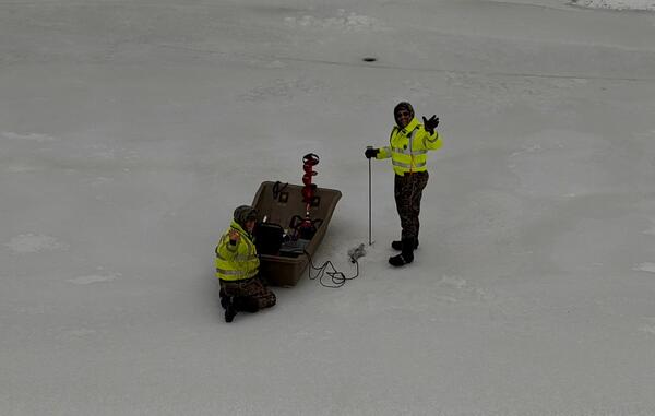 Two scientists next to an ice sled on the frozen Delaware River take a moment to wave at the camera