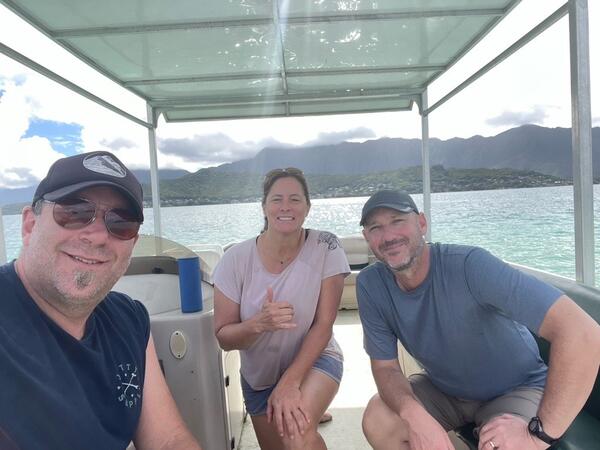 Three smiling people sit in a boat with a lush Pacific Island in the background.