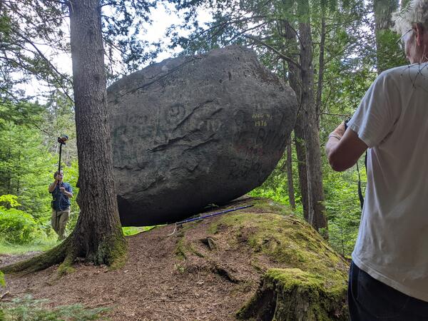 Two scientists on either side of a large boulder perched on bedrock in the forest with scientific instruments.