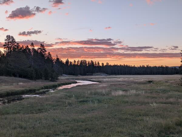 A river winds through a meadow at sunset, with pink clouds in the sky. Trees are in the background.
