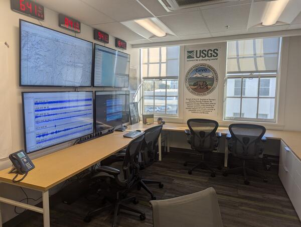 A small room holding a bank of large computer screens, multiple world clocks, and other communications equipment. A desk with multiple rolling chairs wraps around the perimeter of the room.