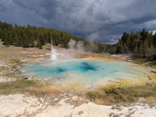 blue-water hot spring with a geyser to one side, forest in background, and storm clouds in the distance