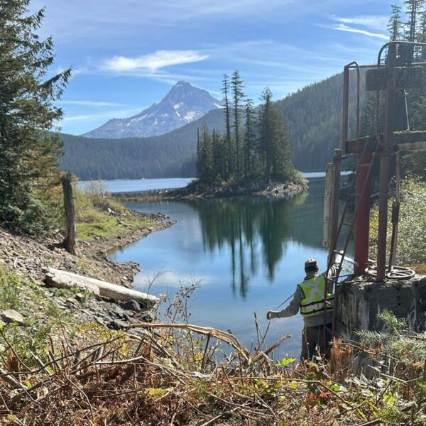 Man lowers a measuring tape into the lake's blue-green water. Island with trees reflect in the water. Mt. Hood in background.