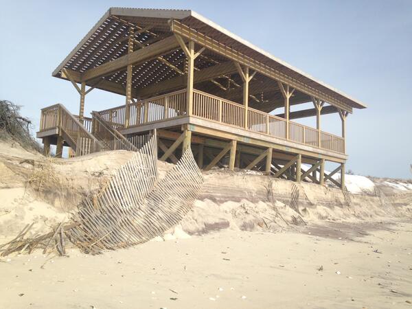 A pavilion on a beach at the edge of an eroded dune is partially covered by sand after a winter storm