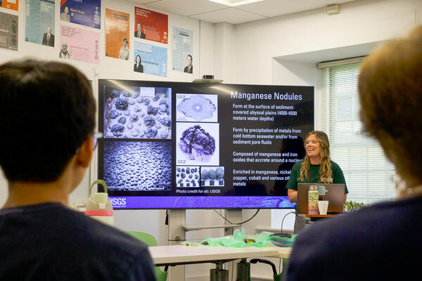 A women stands beside a large presentation screen in front of a classroom.