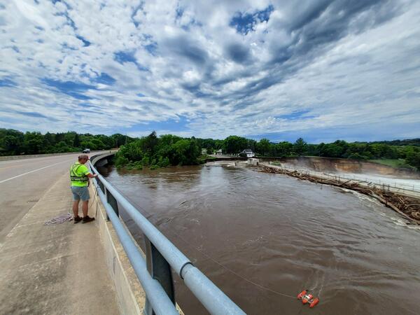 Hydrologic technician in bright green PFD stands on bridge to measure flow with ADCP in water below