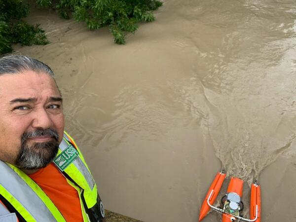 A USGS water expert stands on a bridge measuring floodwaters below, with bright orange specialized monitoring equipment float