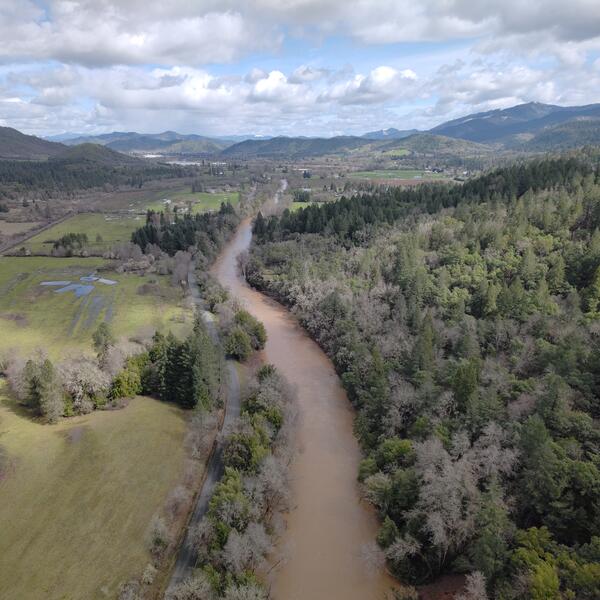 Brown river winds through forested valley with some open pasture on the left. Partly cloudy skies.