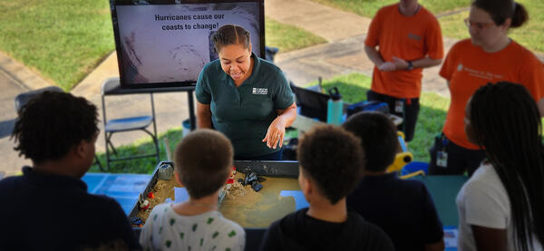 USGS staff at a table with a box filled with sand and water speaks to a group of children