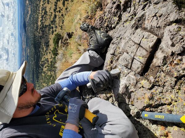 A USGS scientists collects a sample from the surface of a dark-colored rock using a hammer and chisel