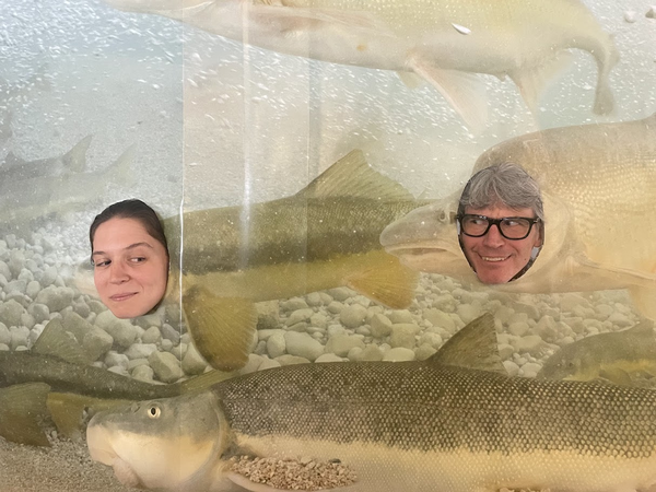 Two USGS scientists pose behind a humpback chub cutout display, their faces framed where the fish’s heads would be.