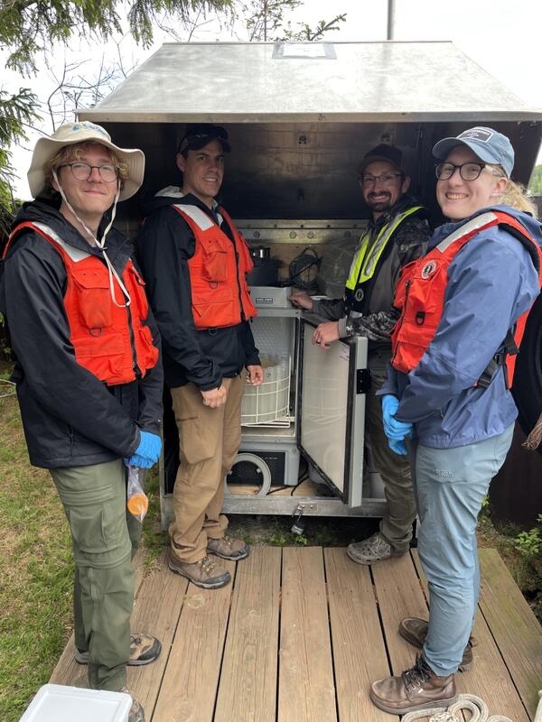 Four smiling scientists stand near an metal housing for an ISCO sampler