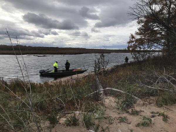 Two people standing in a boat near a sandy shoreline of a slough when other person stands on a the shore