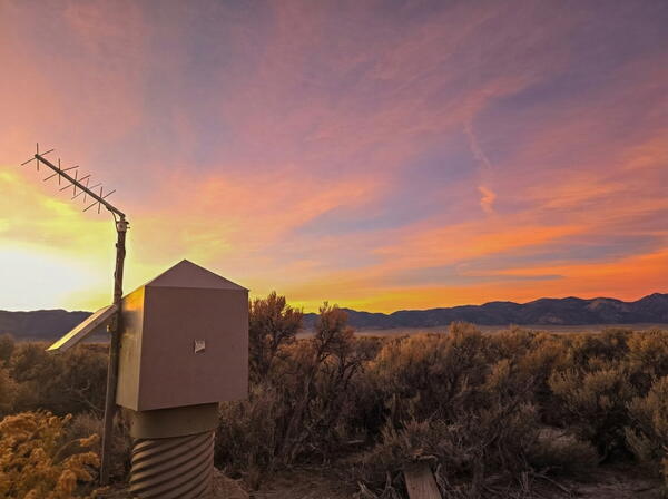 Yellow-orange sunset over desert landscape with groundwater monitoring station and distant mountains.