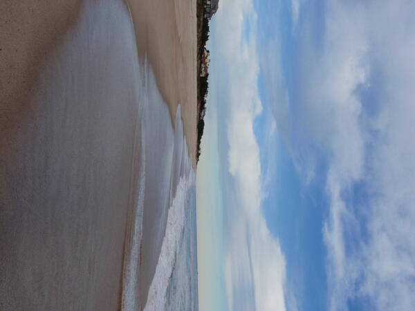 Calm ocean waves on a beach with houses and a cloudy sky in the background