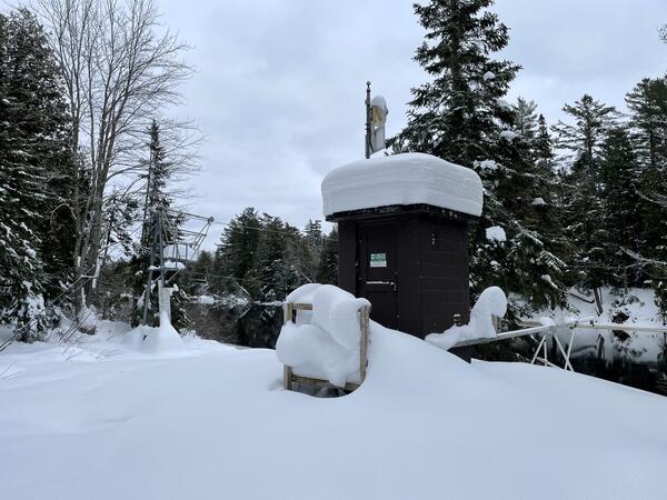 A brown streamgage housing with two feet of fresh snow piled on the roof with a cableway in the background