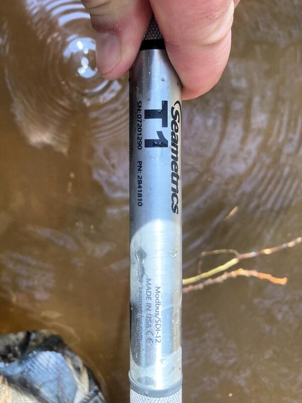 Close up image of hand holding a Seametrics T1 Thermister over brown stream water
