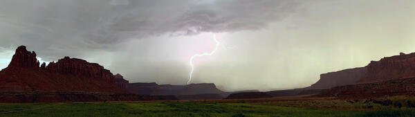 A lightning bolt from a dark and stormy sky strikes a red rock plateau, with green grass fields in the foreground