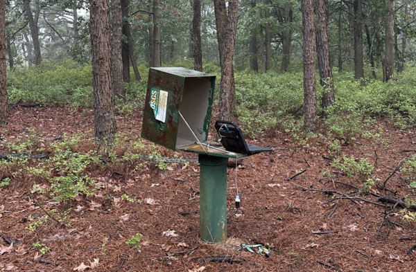 A computer attached to a probe going down a pipe inside a steel box in the woods.