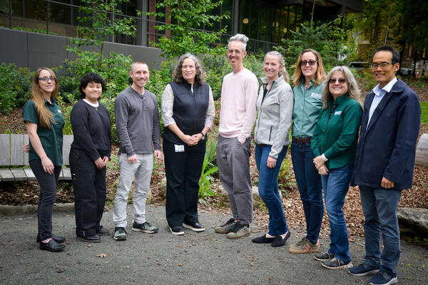 9 people stand in a line with the tallest people in the middle. They are professionally dressed outside amongst greenery