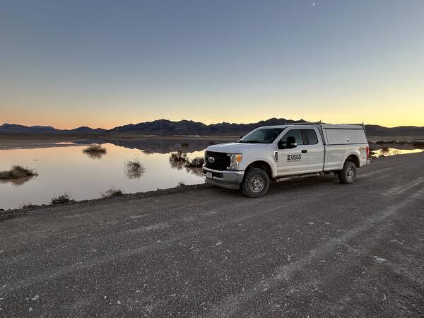 White USGS truck parked on dirt road near shallow-water playa at dusk with silhouetted mountains.