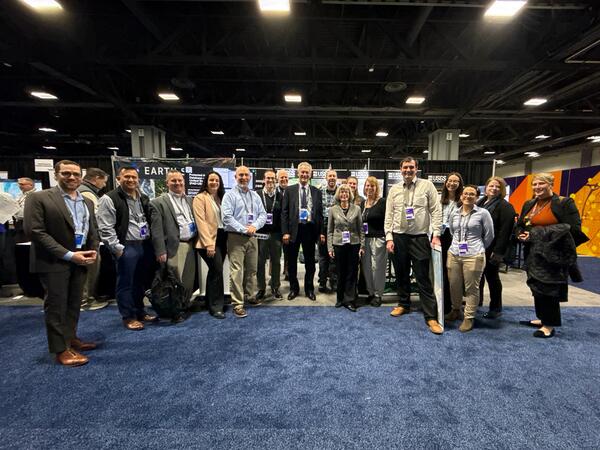 17 people stand in a row at a conference, smiling for the camera