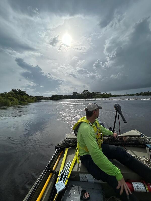 Man in a bright green shirt wearing a baseball cap guides a boat over water 