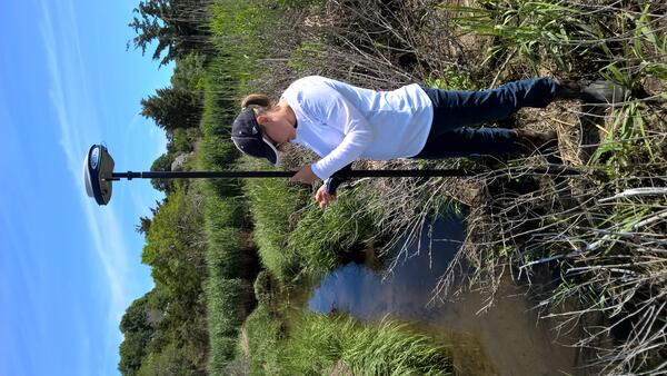 a woman scientist in a baseball hat standing in a marsh holding a long piece of equipment