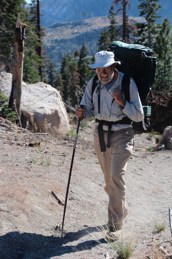 An older geologist wearing khakis, a light blue shirt, and a fisherman's hat hikes up a mountain carrying a large backpack and holding a hiking pole. He is smiling at the photographer.