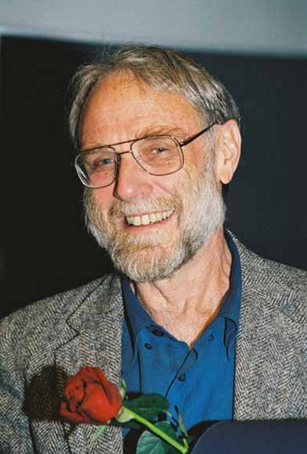 An older man with a salt-and-pepper beard and hair and square glasses, wearing a speckled gray suit and a teal button-down shirt, smiles at the camera. He is holding a red rose. 