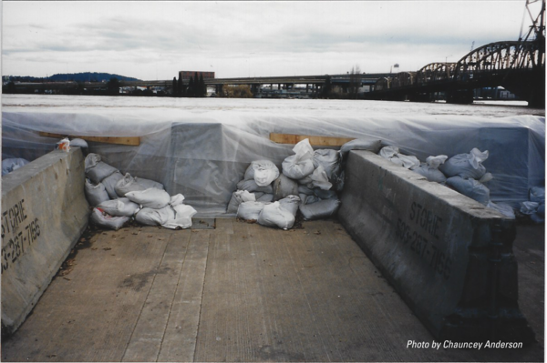 flood waters visible just below the wall supported by concrete blocks and sandbags