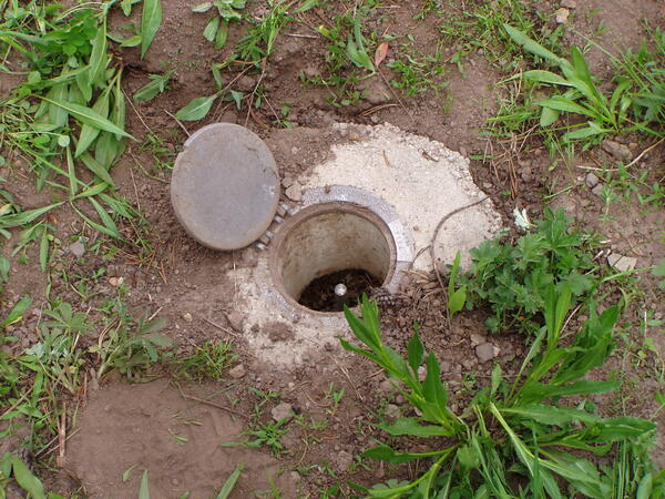 A hinged metal cover, flush with the ground, is open, exposing a metal rod just below the ground surface.