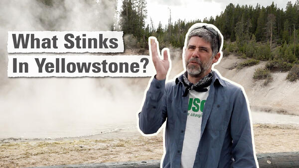 Man in blue shirt waves his hand in front of his face and stands in front of a boiling hot spring