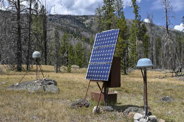 GPS antenna with gray dome on rock in middle ground, solar panels and satellite antenna in foreground, in grassy meadow