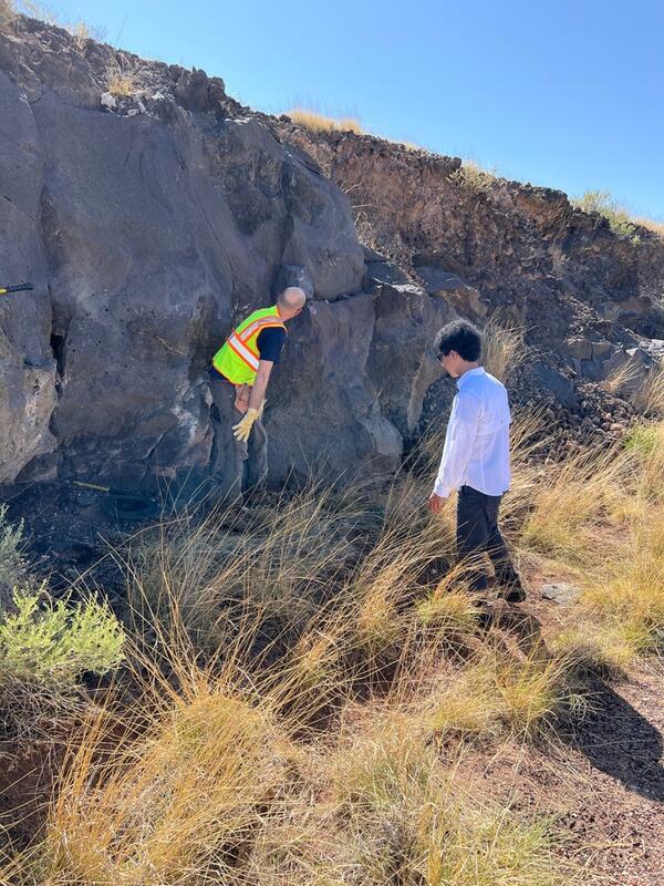 Two men examine a vertical outcrop of gray rock about twice their height, with brown grass at their feet and blue sky above