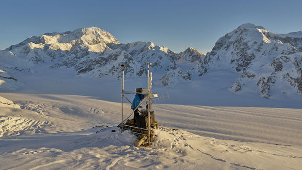 Person in a blue jacket working at a weather station in the snow with mountains in the background 