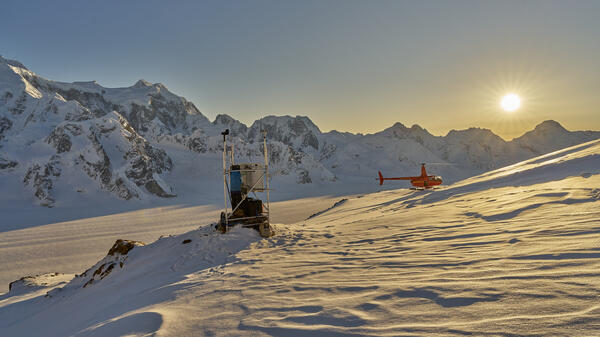 Person in blue jacket at a weather station on snow with the sunshine, mountains, and a red helicopter in the background