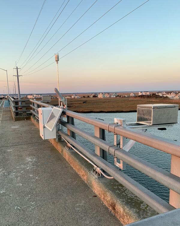 Looking along a bridge rail where a radar unit is suspended out over the river and tied into a control box with a rain gage