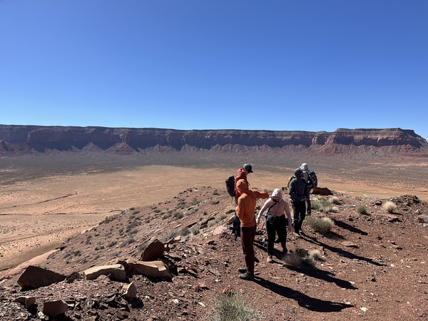 Five people stand in the Utah desert viewing the rocks around them with a large cliff in the background.