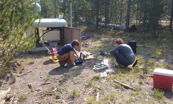 Two people crouching near equipment in a forested clearing and working with hand tools