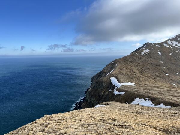 Scenic view of Bearing Sea, blue sky with clouds. Rocky cliff on right side with patches of snow scattered on ground. 