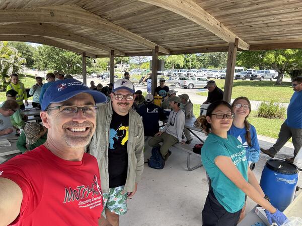 Four people stand in the foreground taking a selfie-style photo, with many people sitting on metal park benches under a large pavilion in the mid-ground, and a road and parking lot in the background